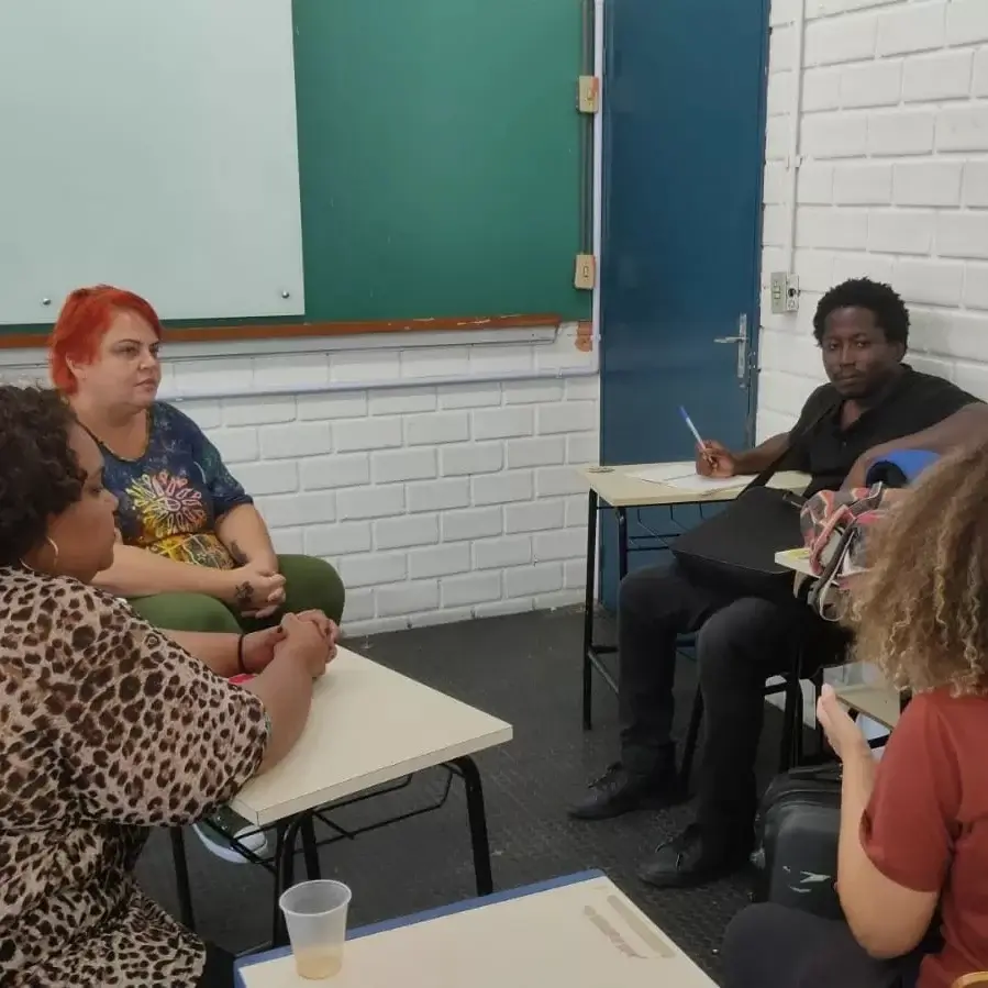 Estudantes conversam sentados nas carteiras de uma sala de aula de paredes de tijolo branco